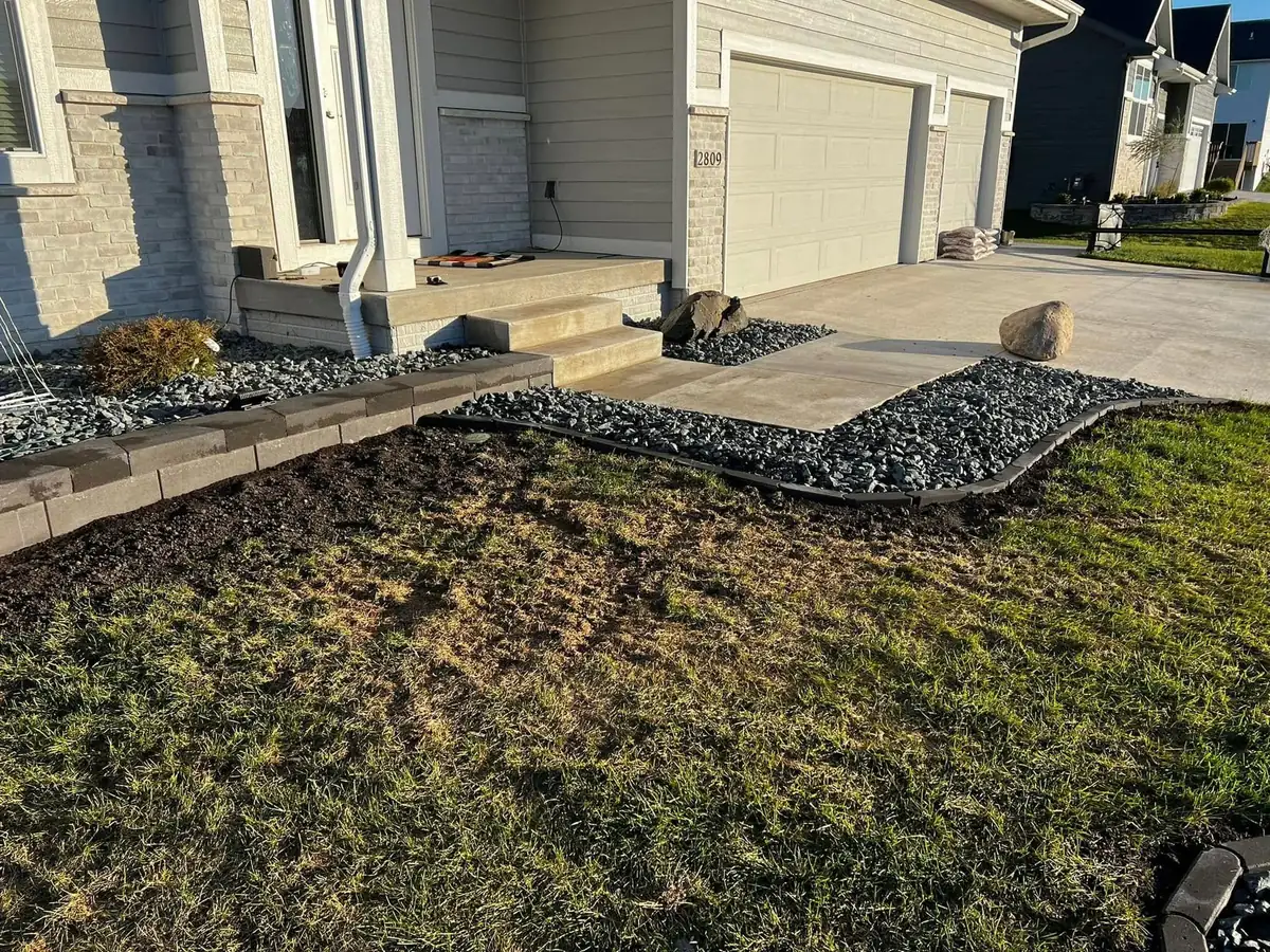 Low segmental block retaining wall and a curved black rock border edging a freshly graded front lawn beside a home's concrete driveway and steps