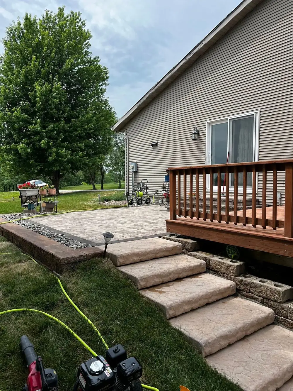 A raised paver patio edged with a block retaining wall and river-rock border, with stone slab steps leading up to a backyard wood deck