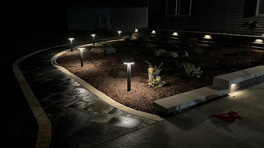 Landscape path lights over a mulched bed beside a stone walkway at night by Larkin Landscape and Design